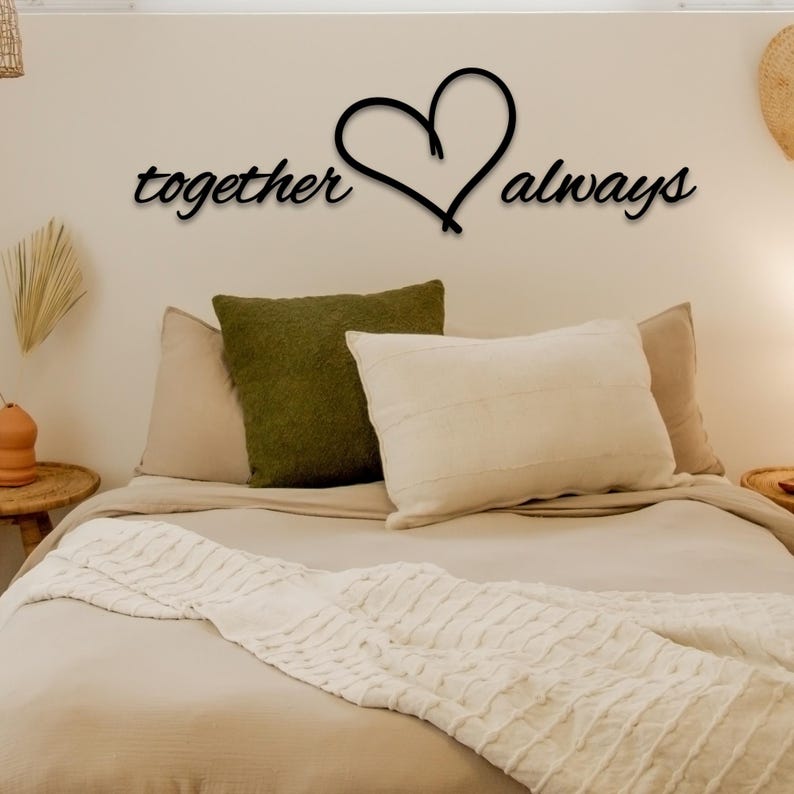 May include: A bedroom with a white bedspread and pillows. The wall above the bed has a black metal sign that says "together always" with a heart in the middle.