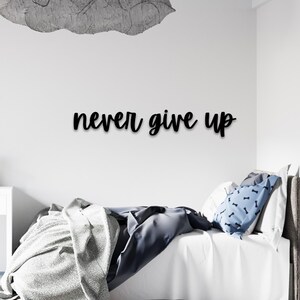 May include: Black cursive text on a white wall reads "never give up." A decorative cloud-shaped element is above the text. A bed with white linens, a blue pillow with a bone pattern, and a gray blanket are in the foreground.