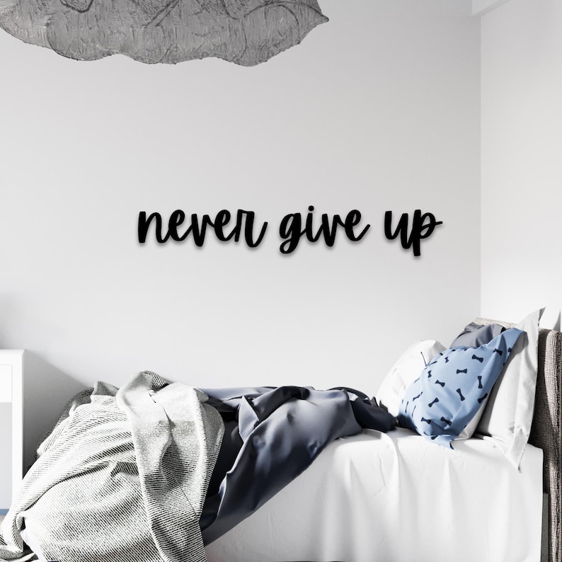 May include: Black cursive text on a white wall reads "never give up." A decorative cloud-shaped element is above the text. A bed with white linens, a blue pillow with a bone pattern, and a gray blanket are in the foreground.