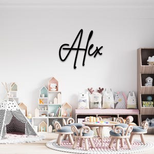May include: A black cursive wooden sign that spells out the name "Alex" is mounted on a white wall. The sign is above a child's playroom with a small table, chairs, and decorative pillows.