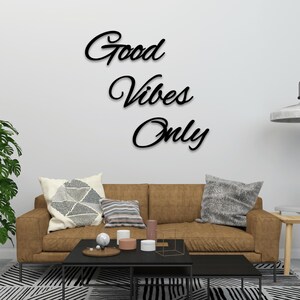 May include: Black cursive wall art that reads "Good Vibes Only" is mounted on a white wall above a brown sofa with decorative pillows. A black coffee table and a patterned rug are in front of the sofa.