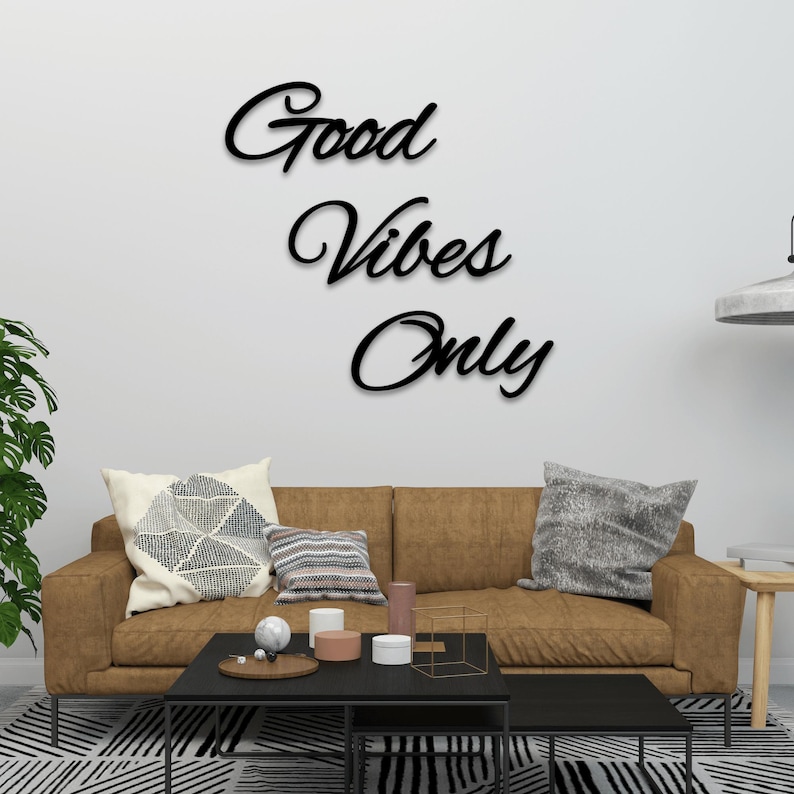 May include: Black cursive wall art that reads "Good Vibes Only" is mounted on a white wall above a brown sofa with decorative pillows. A black coffee table and a patterned rug are in front of the sofa.