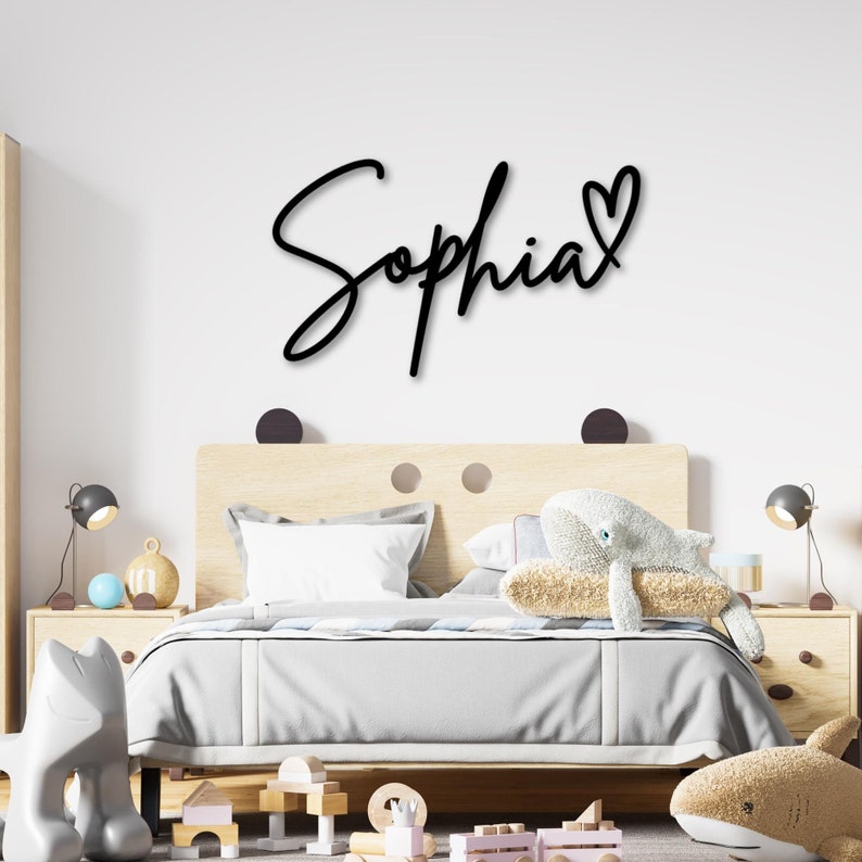 May include: A black wooden sign with the name "Sophia" written in cursive with a heart at the end. The sign is hanging on a white wall above a wooden bed frame with a white comforter and pillows.