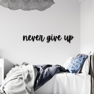 May include: Black wooden wall decor with the text "never give up" in a cursive font. The decor is hanging above a white bed with a gray and white blanket and blue patterned pillows.