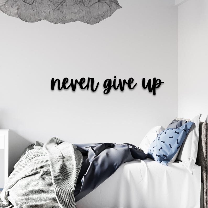 May include: Black wooden wall decor with the text "never give up" in a cursive font. The decor is hanging above a white bed with a gray and white blanket and blue patterned pillows.