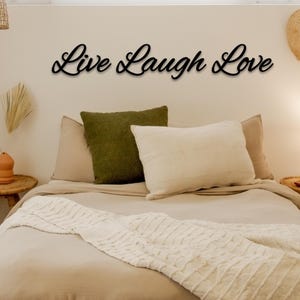 May include: A bedroom with a white bed and a throw blanket. The bed has two pillows, one green and one white. The wall above the bed has a black metal sign that reads "Live Laugh Love".