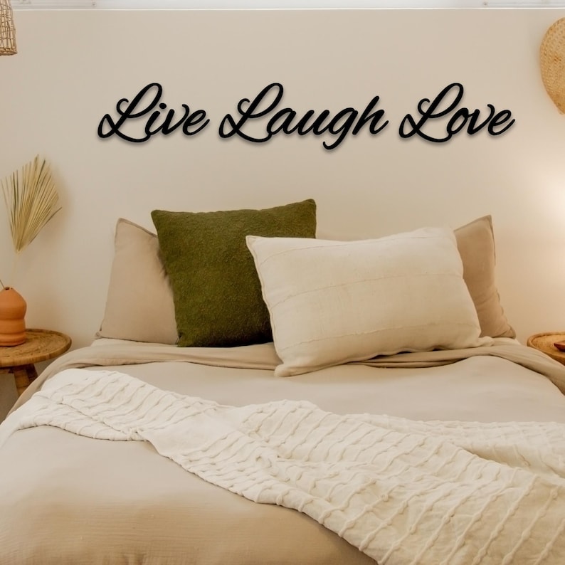 May include: A bedroom with a white bed and a throw blanket. The bed has two pillows, one green and one white. The wall above the bed has a black metal sign that reads "Live Laugh Love".