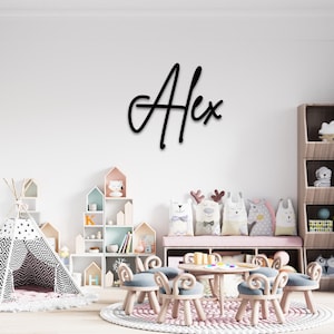 May include: A black wooden name sign with the name "Alex" written in a cursive font. The sign is mounted on a white wall in a child's playroom. The room features a teepee, a bookshelf, and a table with chairs.