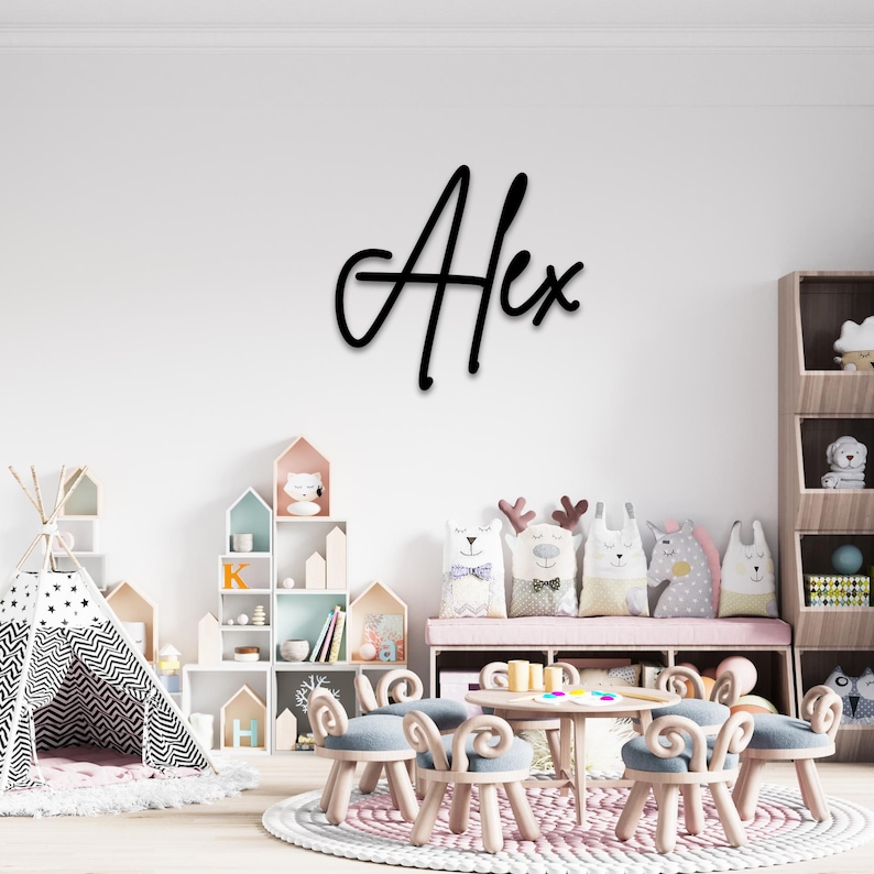 May include: A black wooden name sign with the name "Alex" written in a cursive font. The sign is mounted on a white wall in a child's playroom. The room features a teepee, a bookshelf, and a table with chairs.