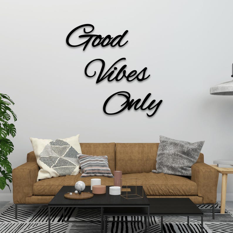 May include: A brown leather sofa with pillows and a black coffee table in front of it. The wall behind the sofa has a black wooden sign that reads "Good Vibes Only".