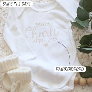 May include: White baby onesie with embroidered text "Baby Charlie April 2023" in gold thread.