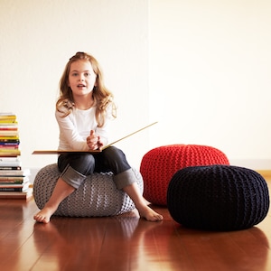 May include: A young girl sits on a gray knitted pouf, holding a book and looking at the camera. There are two other poufs, one red and one black, in the background. A stack of books is on the floor to the left of the girl.