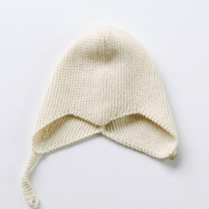 以下が含まれることがあります： クリーム色のニットのベビーボンネット。長いリボンが付いています。ボンネットは柔らかく、質感のある糸でできており、先のとがった形をしています。