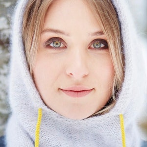 Könnte beinhalten: Nahaufnahme einer Frau, die einen hellgrauen Kapuzen-Schal mit gelben Kordeln trägt. Der Schal ist aus einem weichen, flauschigen Material.