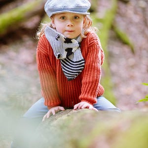 Puede incluir: Un ni&ntilde;o con una gorra de estilo newsboy azul claro, un su&eacute;ter de punto color &oacute;xido y una bufanda gris con un dise&ntilde;o de vaca en blanco y negro. El ni&ntilde;o est&aacute; sentado en un tronco en un entorno forestal.