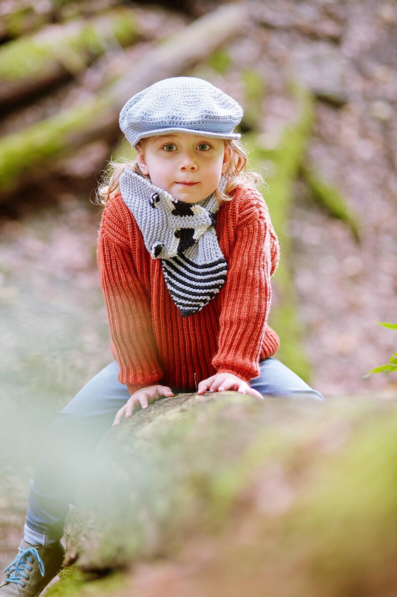 Puede incluir: Un ni&ntilde;o con una gorra de estilo newsboy azul claro, un su&eacute;ter de punto color &oacute;xido y una bufanda gris con un dise&ntilde;o de vaca en blanco y negro. El ni&ntilde;o est&aacute; sentado en un tronco en un entorno forestal.