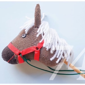 Könnte beinhalten: Ein brauner gestrickter Pferdekopf mit rotem Zaumzeug und weißer Garnmähne. Der Pferdekopf ist an einem Holzstab mit grünen Schnurzügeln befestigt.