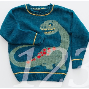 Op de afbeelding: Teal gebreide trui met een groen dinosaurusontwerp met rode en roze accenten. De trui heeft gele biezen rond de halslijn, manchetten en zoom. Het nummer "12" is gedeeltelijk zichtbaar.
