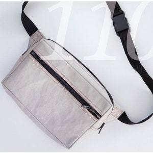 Könnte beinhalten: Eine graue Bauchtasche mit einem schwarzen Reißverschluss und einem schwarzen verstellbaren Riemen. Die Tasche hat ein rotes Stichdetail um den Reißverschluss.