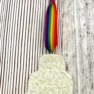 May include: A cream-colored, textured ornament with a rainbow ribbon and a rainbow stripe. The ornament is shaped like a layered cake. The ribbon is tied at the top and is hanging down.