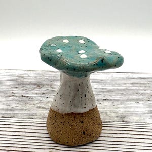 May include: A ceramic mushroom sculpture featuring a glossy teal cap with white speckled dots and a smooth white midsection. The base is made of unglazed, textured brown clay with dark speckles, creating a distinct contrast in colour and surface finish.