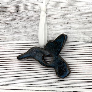 May include: A ceramic hummingbird ornament with a heart-shaped cutout. The bird is glazed in shades of blue and brown, with a white ribbon for hanging. The ornament is set against a white and gray striped background.