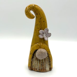 May include: A ceramic gnome figurine featuring a tall, curved mustard yellow hat with dark speckles. A light pink five-petaled flower is attached to the side of the hat. The gnome has a round, speckled light pink nose and a textured, tan-colored beard.