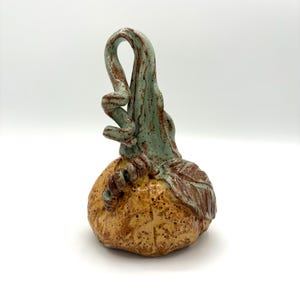 May include: A ceramic sculpture shaped like a pumpkin with a textured, speckled tan body and a tall, curved, light green stem. The stem features a spiraling tendril and a single leaf detail, with brown speckles throughout the entire piece.