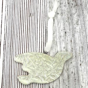 May include: A pale green ceramic ornament shaped like a bird in flight, featuring a textured surface with a glossy finish. The ornament includes a white fabric ribbon tied in a knot at the top for hanging, providing a simple and functional design.