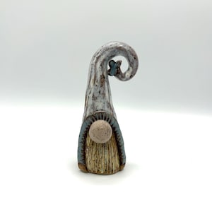 May include: A ceramic gnome figurine featuring a tall, curled hat in mottled white and brown tones with a small blue butterfly accent. The gnome has a round, speckled nose and a textured, tan-coloured beard with vertical brown lines.