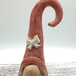 Op de afbeelding: Een keramieken kabouterfiguur met een hoge, getextureerde roze muts die aan de bovenkant omkrult. De muts is versierd met een kleine, witte, gebeeldhouwde strik. De kabouter heeft een gladde, ronde, lichtgekleurde neus en een lange, verticaal gegroefde witte baard.