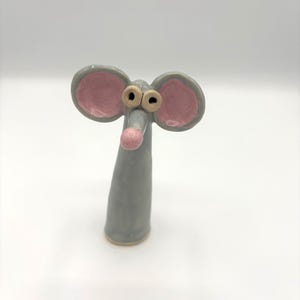 May include: A whimsical ceramic mouse figurine. The mouse has large, round ears with pink interiors, small brown eyes, and a pink nose. The body is a light gray color, and the figurine has a simple, cartoonish design.