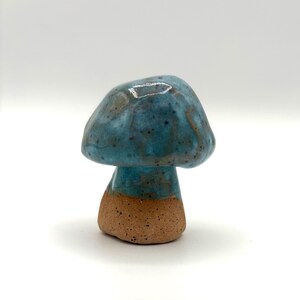 May include: A small ceramic mushroom figurine featuring a glossy, speckled blue glaze on the rounded cap and upper stem, transitioning into a textured, unglazed, earthy brown clay base that provides a stable foundation for the piece.