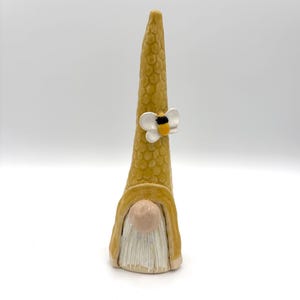 Puede incluir: Una figura alta de gnomo de cerámica con un sombrero puntiagudo amarillo de textura de panal, adornado con un pequeño detalle de abeja blanca y amarilla. El gnomo tiene una nariz redonda de color rosa pálido y una barba blanca texturizada.