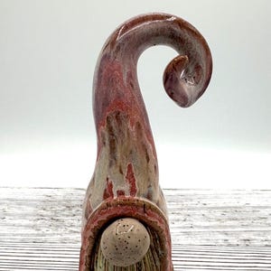 May include: A ceramic gnome figurine featuring a tall, curved, pointed hat with a glossy, mottled red and beige glaze. The gnome has a round, speckled beige nose and a textured, vertically grooved beard in shades of brown and cream.