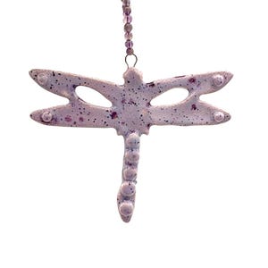 Op de afbeelding: Een lavendelkleurig libelle-ornament met gespikkelde details en een kralenreeks om op te hangen. De libelle heeft opengewerkte vleugels en een gesegmenteerd lichaam. Het ornament is ontworpen om op te hangen en voegt een vleugje natuurlijke decoratie toe.