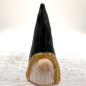 May include: A ceramic gnome figurine with a dark green, speckled hat and a tan, textured beard and hat trim. The gnome has a round, light pink nose and a white beard. The figurine is on a white and brown striped surface.