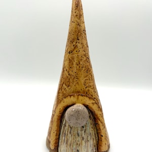 May include: A tall, conical ceramic gnome figurine featuring a speckled tan and brown glaze. The piece has a prominent, smooth, light-colored round nose positioned above a textured, vertically grooved beard in shades of cream and brown.
