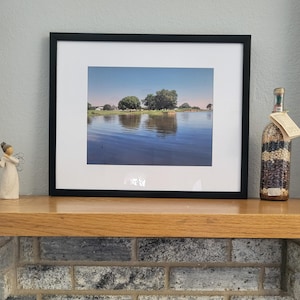 May include: A framed photograph of a lake with trees reflected in the water. The lake is a light blue colour and the trees are green. The frame is black.