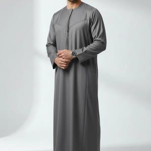 May include: A gray thobe, a traditional Arab garment, with long sleeves and a mandarin collar. The thobe features a decorative stitched design along the front and a side pocket. The person is wearing a silver watch and black shoes.