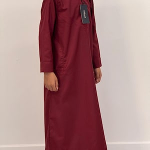 May include: A long-sleeved, burgundy-colored garment with a mandarin collar and a small pocket. The garment has a tag that reads "ANDAAZ". The fabric appears to be a lightweight cotton or linen blend. The garment is floor-length.