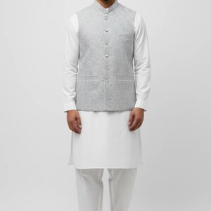 May include: A light grey sleeveless waistcoat with a subtle tonal floral pattern, featuring a mandarin collar and a front button closure, worn over a long-sleeved white tunic and white trousers, paired with brown pointed-toe slip-on shoes.