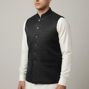 May include: A black waistcoat with a subtle geometric pattern. The vest has a mandarin collar and is buttoned down the front. It is worn over a white long-sleeved shirt and paired with white pants. The overall style is formal.