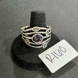May include: Silver-toned ring with an oval purple gemstone and smaller accent stones. The ring has a multi-layered design with a textured band. The ring is displayed on a black ring holder.