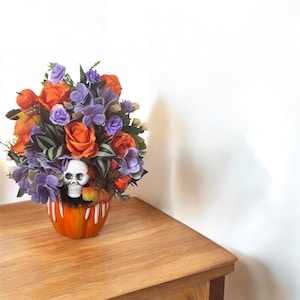 May include: A Halloween floral arrangement in an orange pumpkin-shaped vase. The arrangement features orange roses, purple hydrangeas, and a white skull. The vase sits on a wooden surface, creating a festive and decorative display.