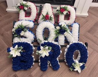 May include: A floral tribute display featuring three rows of letters spelling DAD, SON, and BRO. The letters are covered in white and dark blue carnations, accented with white lilies, dark blue roses, and pink roses, all arranged on a metal stand.