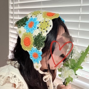 May include: A colorful, crocheted head covering featuring a floral design. The head covering has a cream-colored base with blue, orange, and green flower accents. The head covering is tied with cream-colored ties.