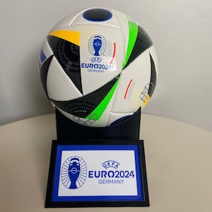 May include: A white and black soccer ball with green and yellow accents. The ball has the UEFA EURO2024 logo and the text "Germany". The ball is on a black stand with a blue and white plaque that reads "UEFA EURO2024 GERMANY".