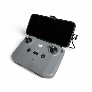 May include: A grey DJI drone remote control with a black phone holder attached. The remote has a power button, a joystick, and a screen that displays the drone's status.