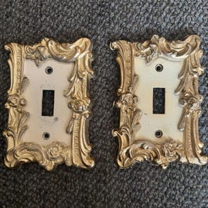 Puede incluir: Dos cubiertas de interruptores de luz ornamentadas de estilo vintage. Cada cubierta tiene una abertura rectangular para el interruptor y está rodeada por un marco dorado detallado con volutas. El fondo es una superficie gris texturizada.